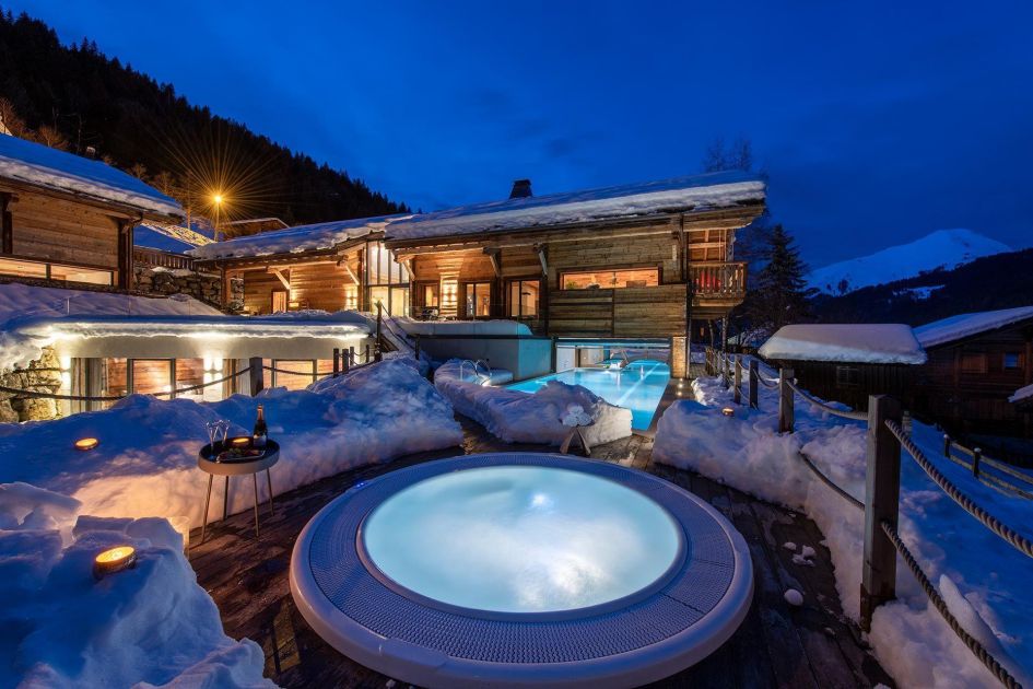 Chalet Joux Plane's outdoor hot tub and indoor-outdoor pool pictured in front of the chalet under snow and blue night's sky.