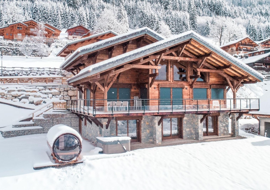 Chalet Le Whymper's stone and wooden exterior covered in snow, with an outdoor hot tub and barrel sauna also seen to the left of the image.