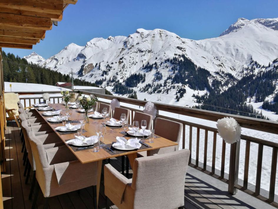 Outdoor dining at UberHaus with a large table on the sunny terrace and a snowy mountain view. 