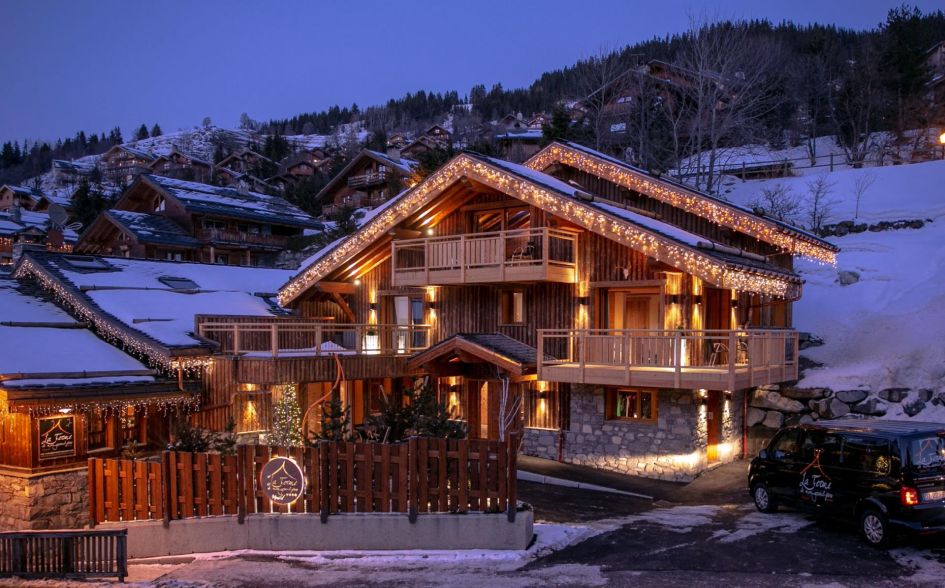 The exterior of Ferme Mon Grand Pere, one of the top luxury chalets in Méribel, lit by twinkling fairy lights to show its stone and wooden exterior.