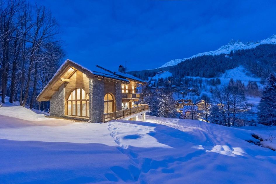 Chalet Alya sits amongst snow and forest, illuminated by light to show its stone and wooden exterior.