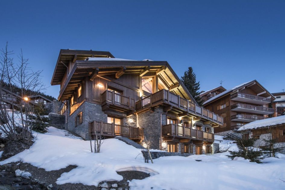 Chalet Le Grenier's dark wooden and stone exterior sits behind snow and under evening blue skies.