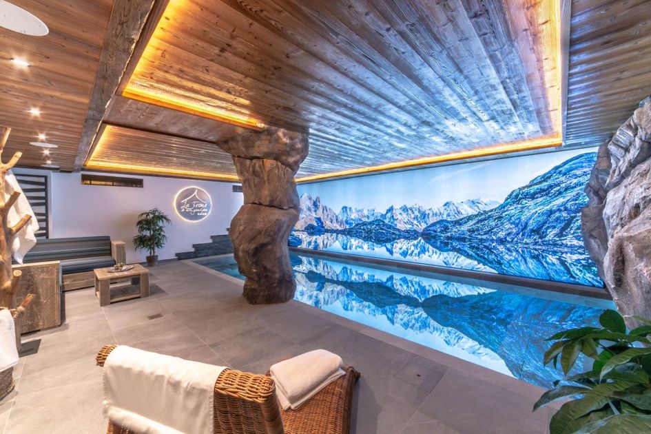 The indoor swimming pool of Ferme Mon Grand Pere. A lit panoramic mountain picture spreads against the back wall, under wooden cladded ceilings.