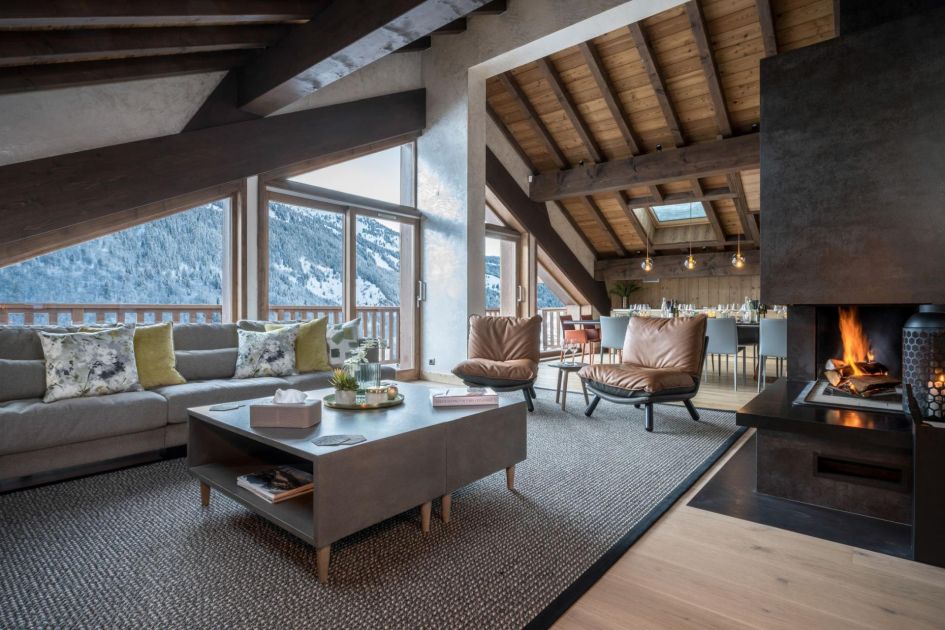 The living area of Chalet Serendipity featuring a wood-burning fireplace, grey furnishings and slanted wooden ceiling with views out to snowy mountains outside.