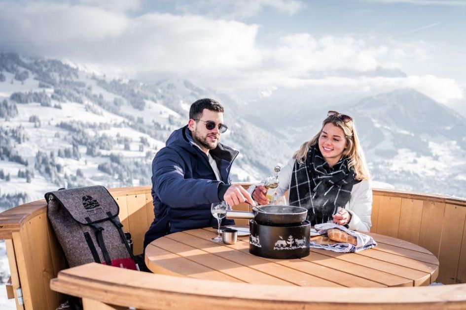 Man and woman enjoying a bottle of wine and fondue at a table in a mountain restaurant in Gstaad, with the snowy landscape behind them.