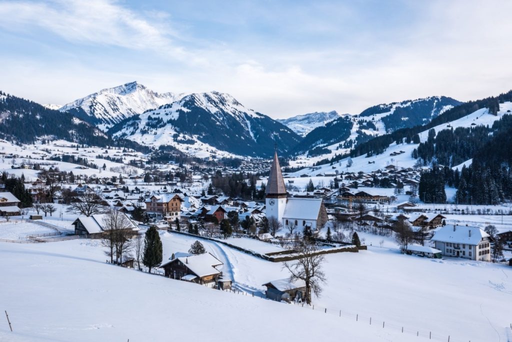 Picturesque ski village Gstaad Saanen with snow covered chalet roofs and a quintessential church occupying the foreground and tree-lined snowy mountains in the background. 